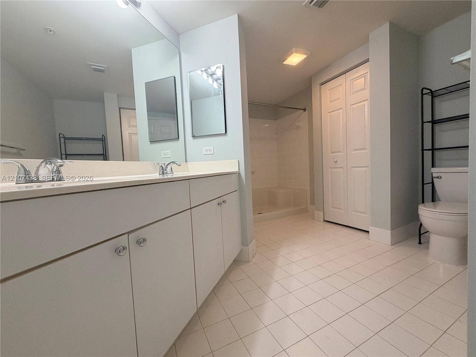 Bathroom featuring a dual-sink vanity with solid-surface countertop, full-width wall mirror, walk-in shower enclosure, and white ceramic tile flooring