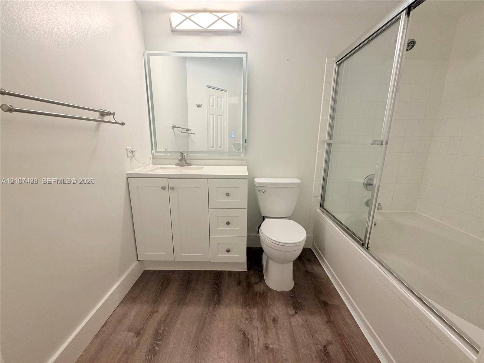White vanity with a rectangular sink, illuminated mirror, wood-finish flooring, and a shower-tub combination with glass sliding doors