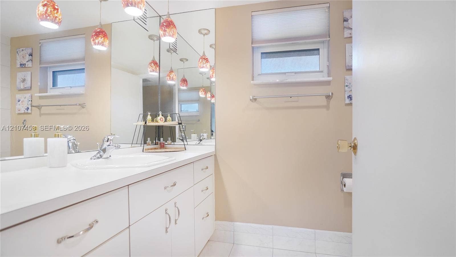111 N Pompano Beach Boulevard, #304, Pompano Beach, FL 33062 White vanity with integrated countertop and sink, large wall-to-wall mirror, multi-pendant lighting, tile flooring, and a window with a built-in shade
