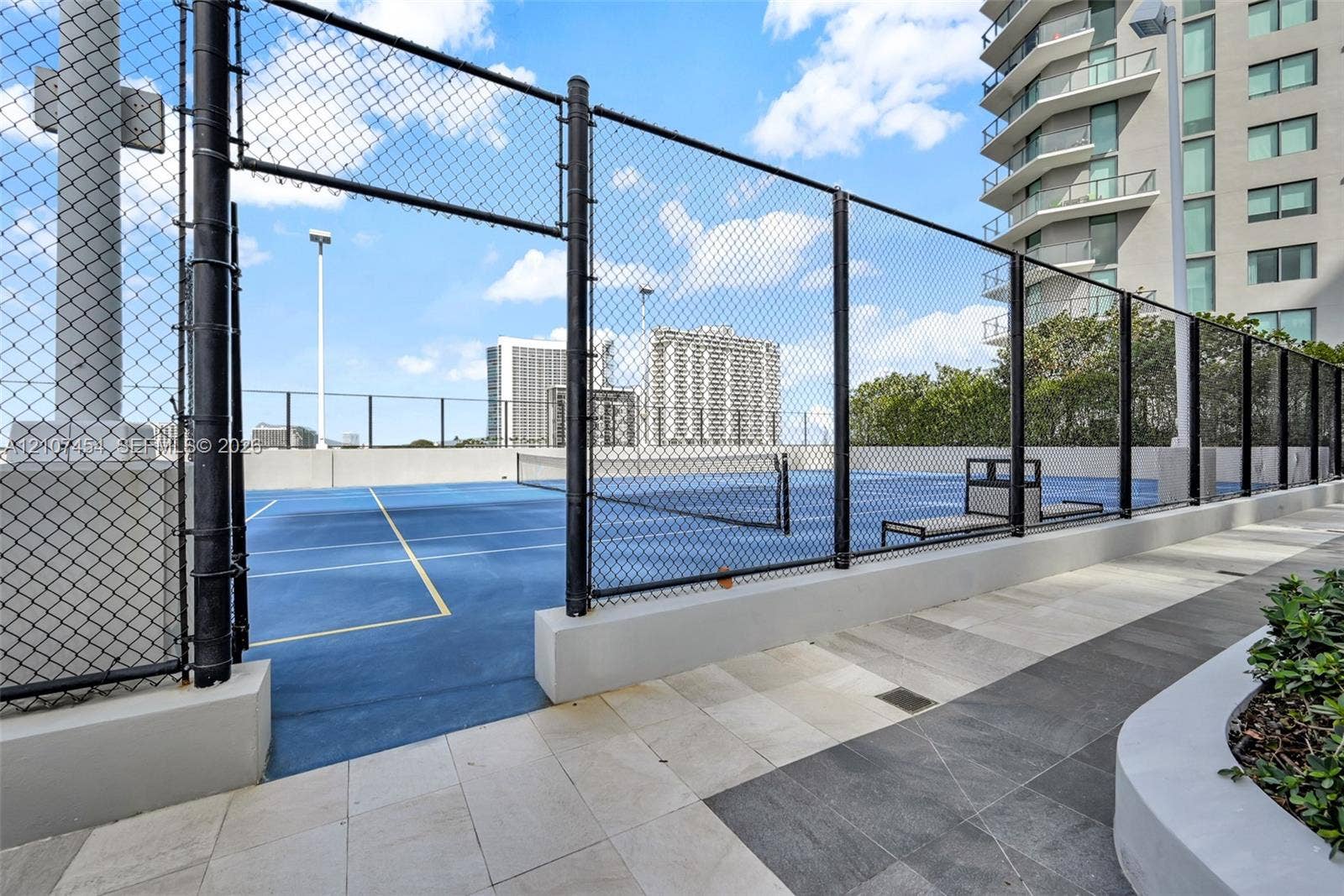 501 NE 31st Street, #1906, Miami, FL 33137 Tennis court featuring a blue hard surface, perimeter chain-link fencing, built-in court seating, and stone-tiled walking paths