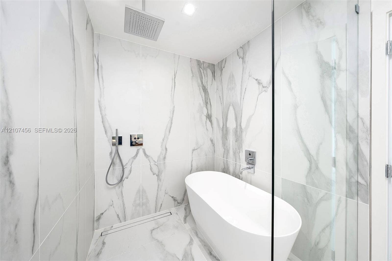 1400 Biscayne Boulevard, #2604, Miami, Miami, FL 33132 Wet room featuring floor-to-ceiling veined stone-style tile, a freestanding soaking tub, a ceiling-mounted rainfall showerhead, a wall-mounted hand-held sprayer, and a glass partition