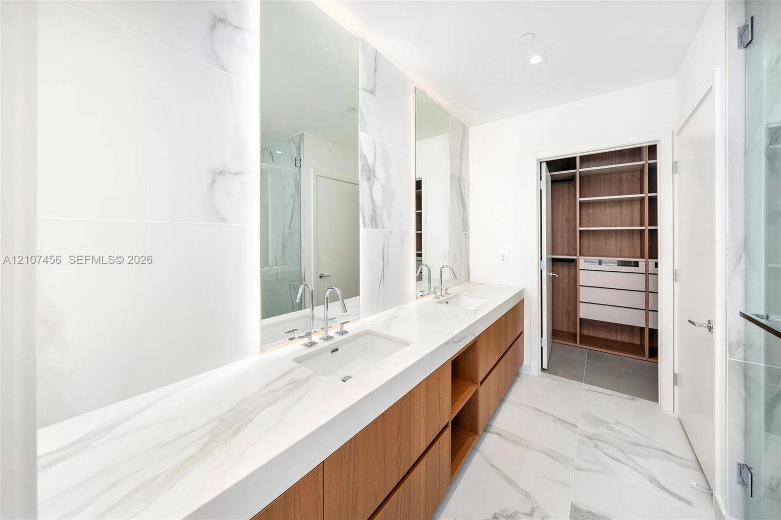 1400 Biscayne Boulevard, #2604, Miami, Miami, FL 33132 Dual-sink vanity featuring a stone-composite countertop, wood-finish cabinetry, floor-to-ceiling porcelain tile wall cladding, integrated vanity mirrors with LED lighting, and custom built-in closet shelving
