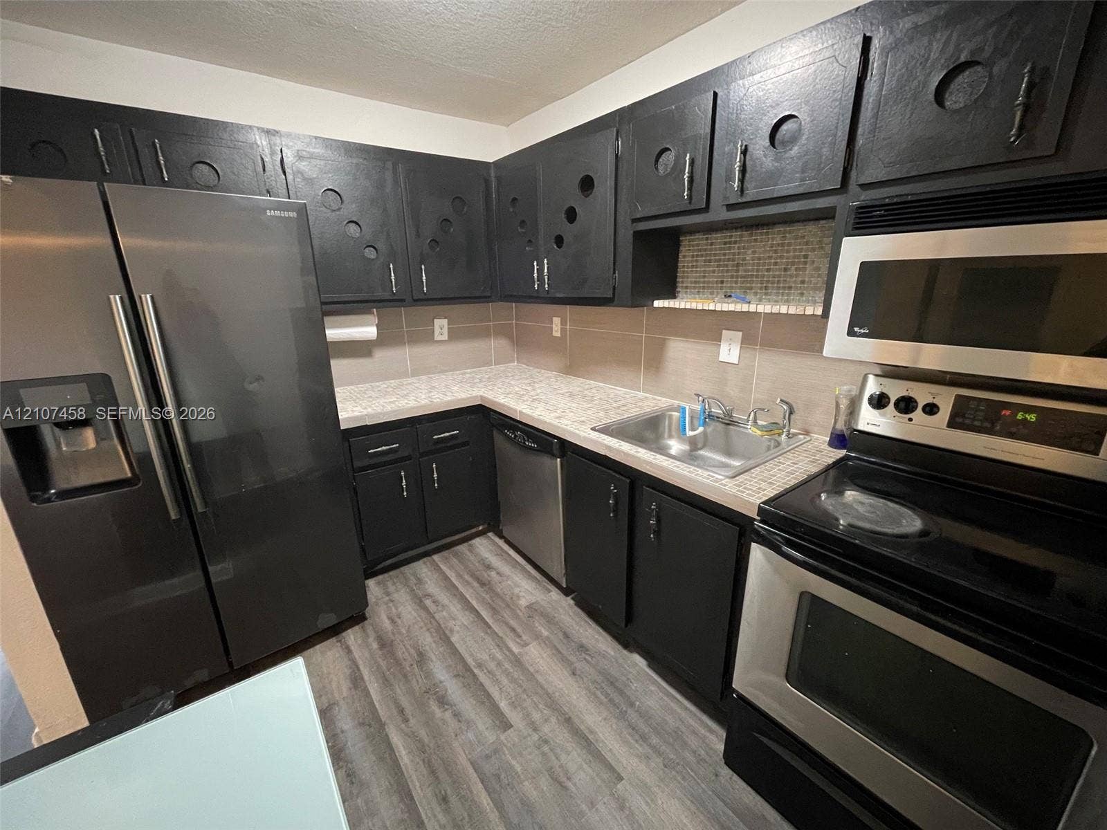 7300 NW 17th Street, #421, Plantation, Plantation, FL 33313 Kitchen featuring custom black cabinetry with circular detailing, stainless steel appliances, patterned laminate countertops, glass tile backsplash, and wood-finish flooring