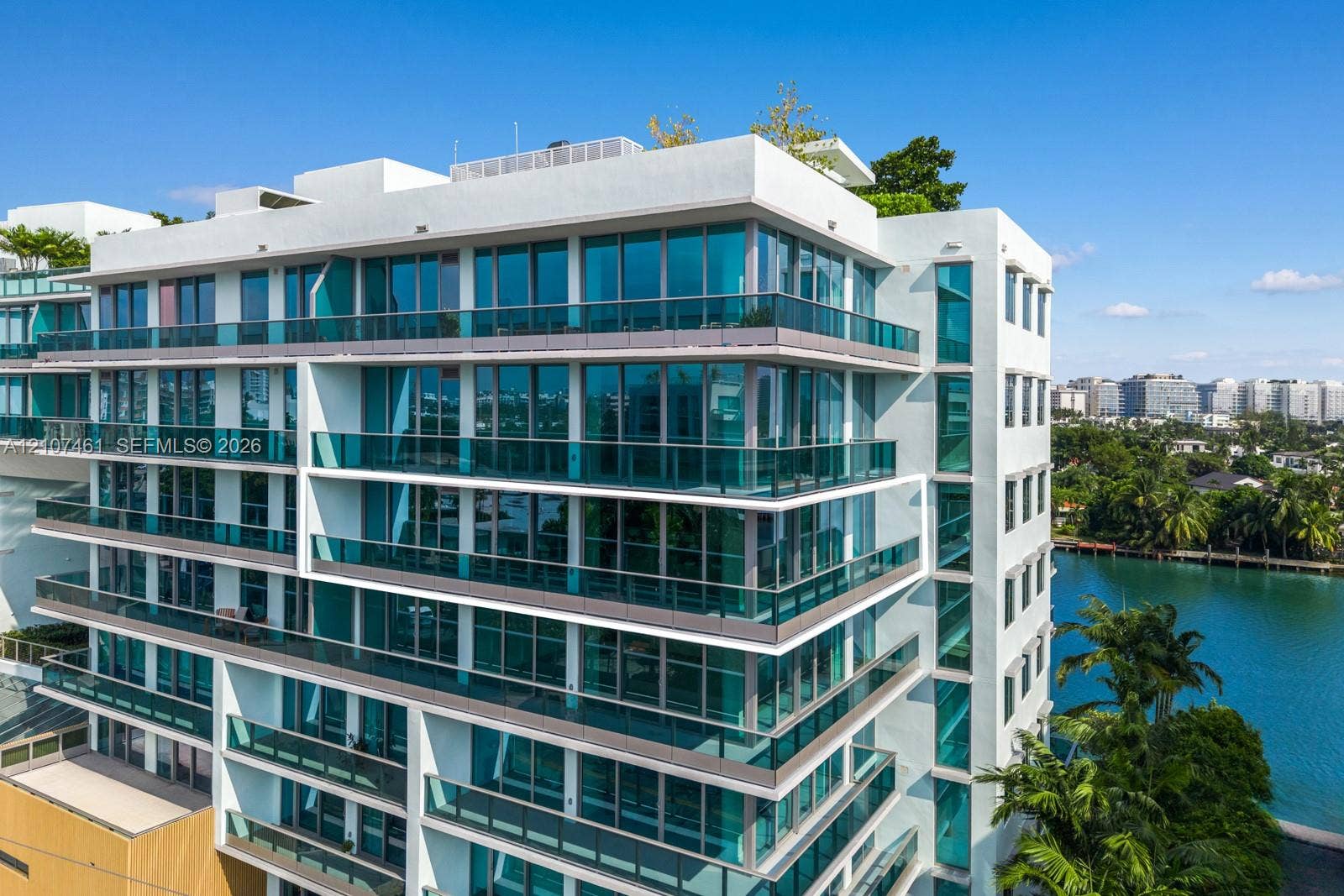 9201 E Bay Harbor Drive, #602, Bal Harbour, FL 33141 Modern multi-story building featuring white stucco facade, expansive floor-to-ceiling glass paneling, tiered balconies with tinted glass railings, and a roof terrace