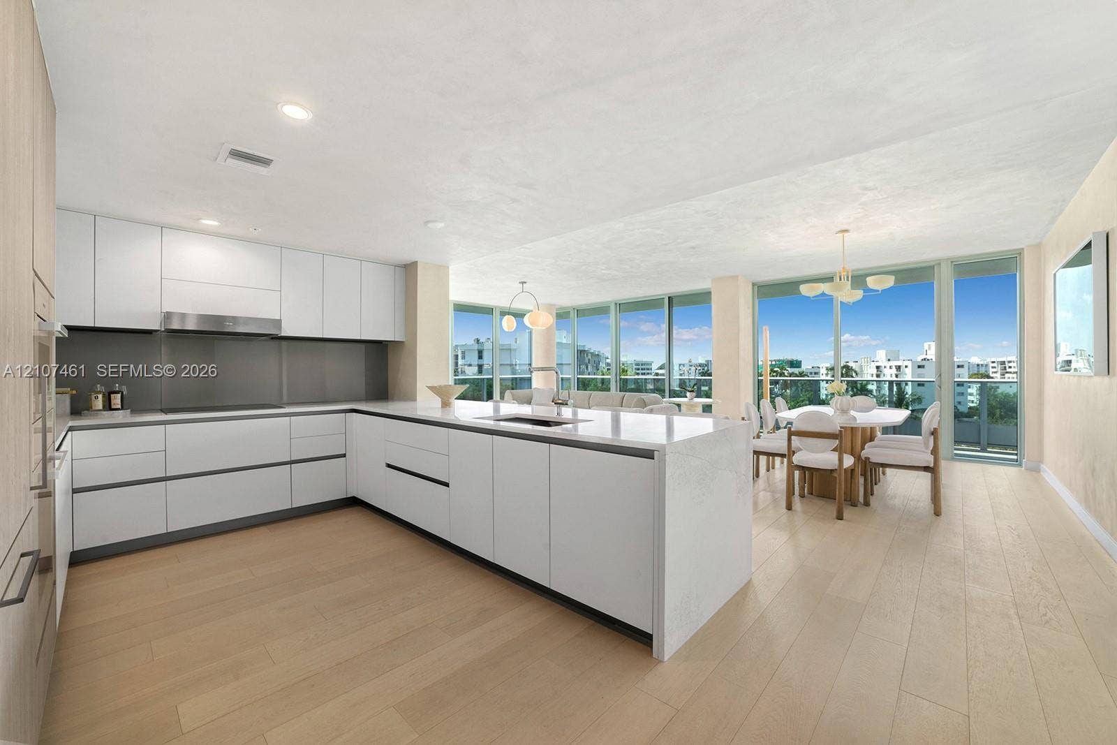 Contemporary kitchen featuring white handleless cabinetry, a substantial quartz-topped center island with integrated sink, stainless steel range hood, and light-toned wood-finish flooring, complemented by floor-to-ceiling glass wall panels