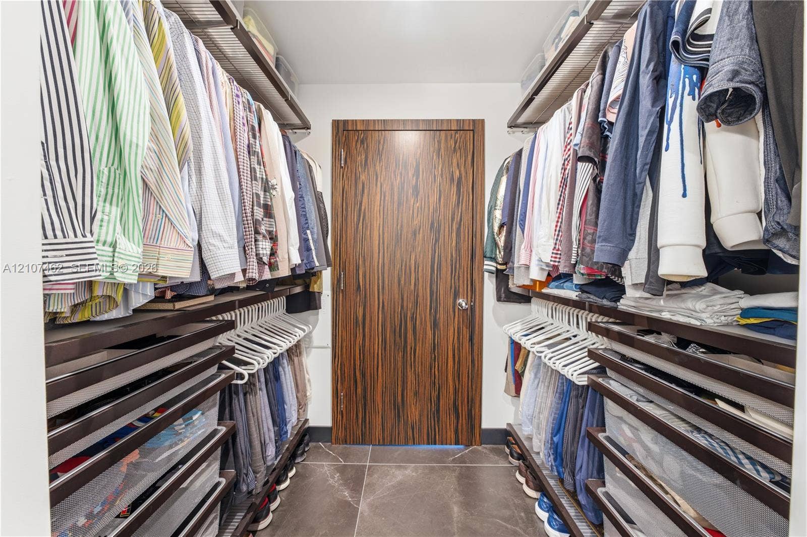 455 NE 39th Street, #110, Miami, FL 33137 Walk-in closet featuring dual-sided integrated shelving systems, multi-tier hanging rods, pull-out storage drawers, and large-format tile flooring with a wood-finish interior door