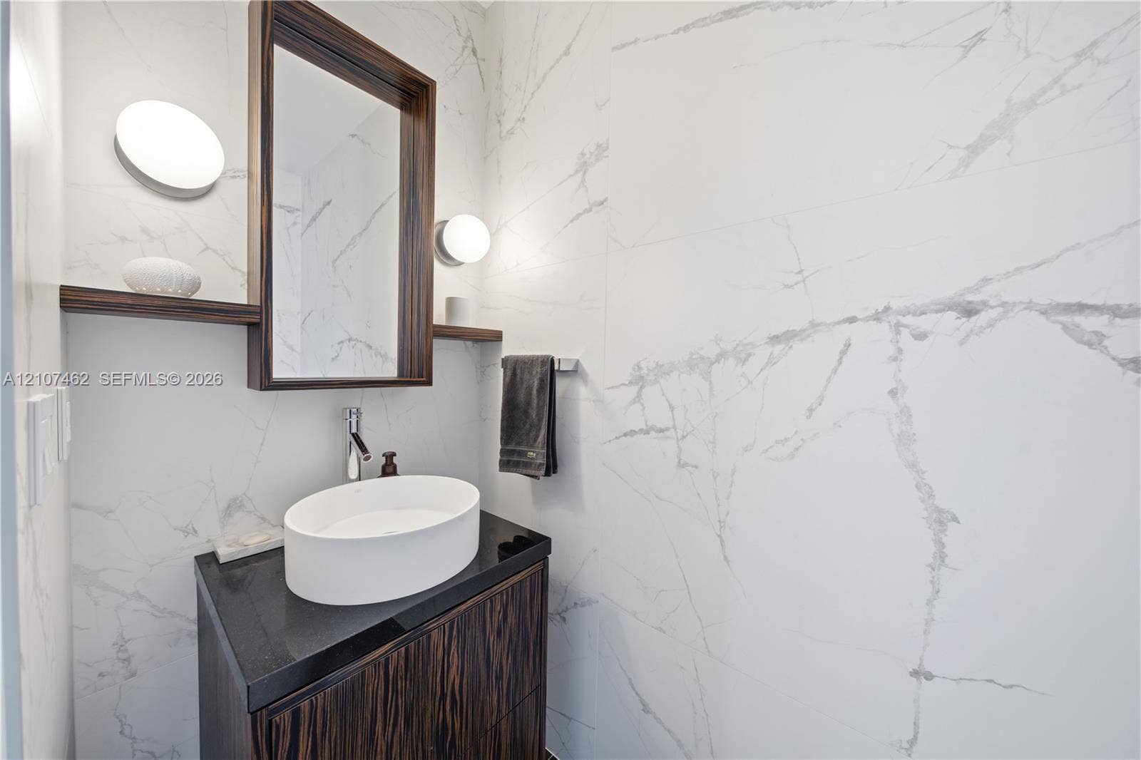 455 NE 39th Street, #110, Miami, FL 33137 Dark wood-finish vanity with a stone countertop, white oval vessel sink, wall-mounted mirror with integrated shelving, and floor-to-ceiling stone-style wall tile