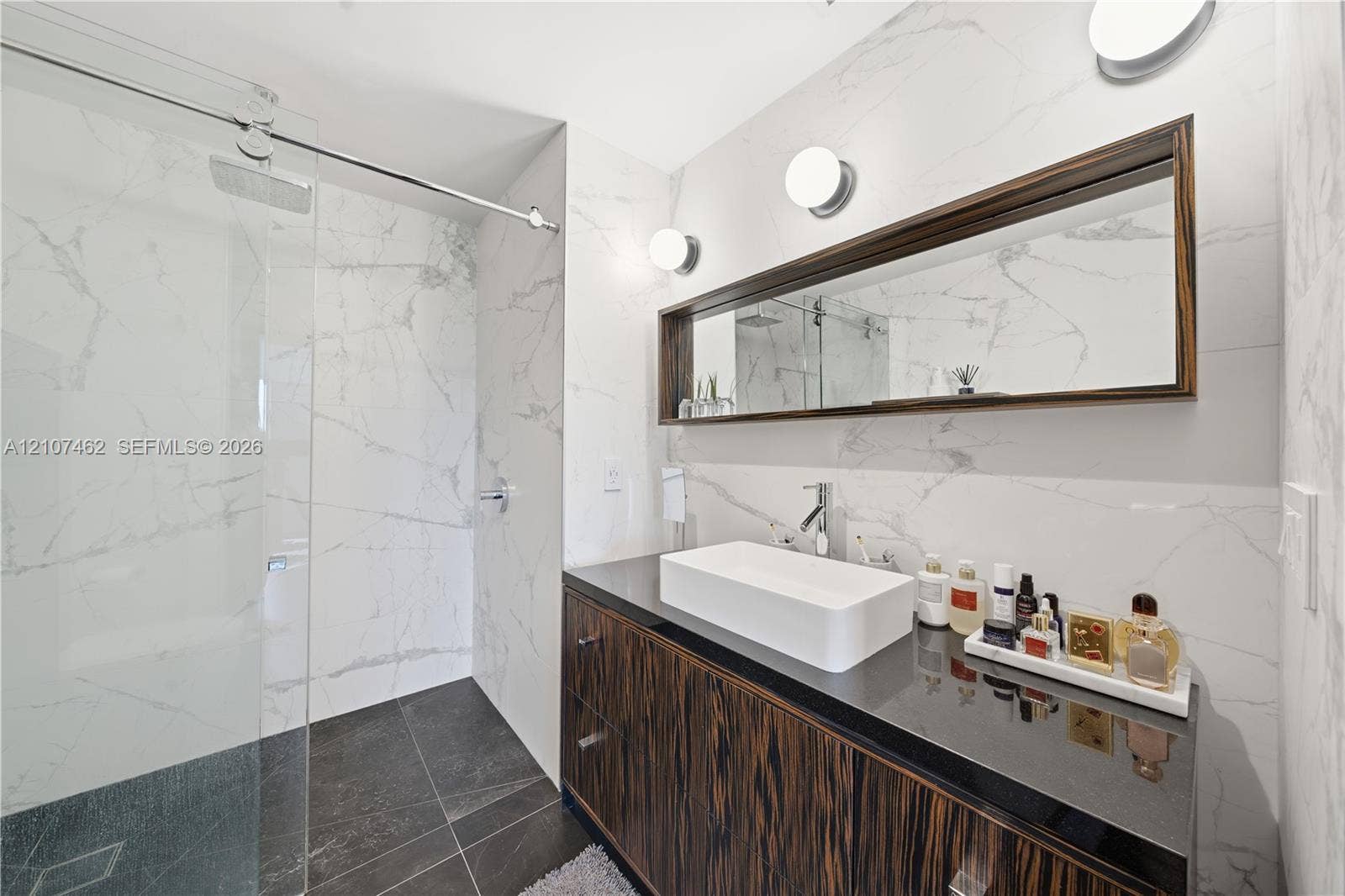 455 NE 39th Street, #110, Miami, FL 33137 Bathroom featuring a wall-mounted vanity with a dark wood-finish cabinet and dark stone countertop, rectangular vessel sink, floor-to-ceiling large-format tile surround, frameless glass shower enclosure, and integrated mirror cabinetry