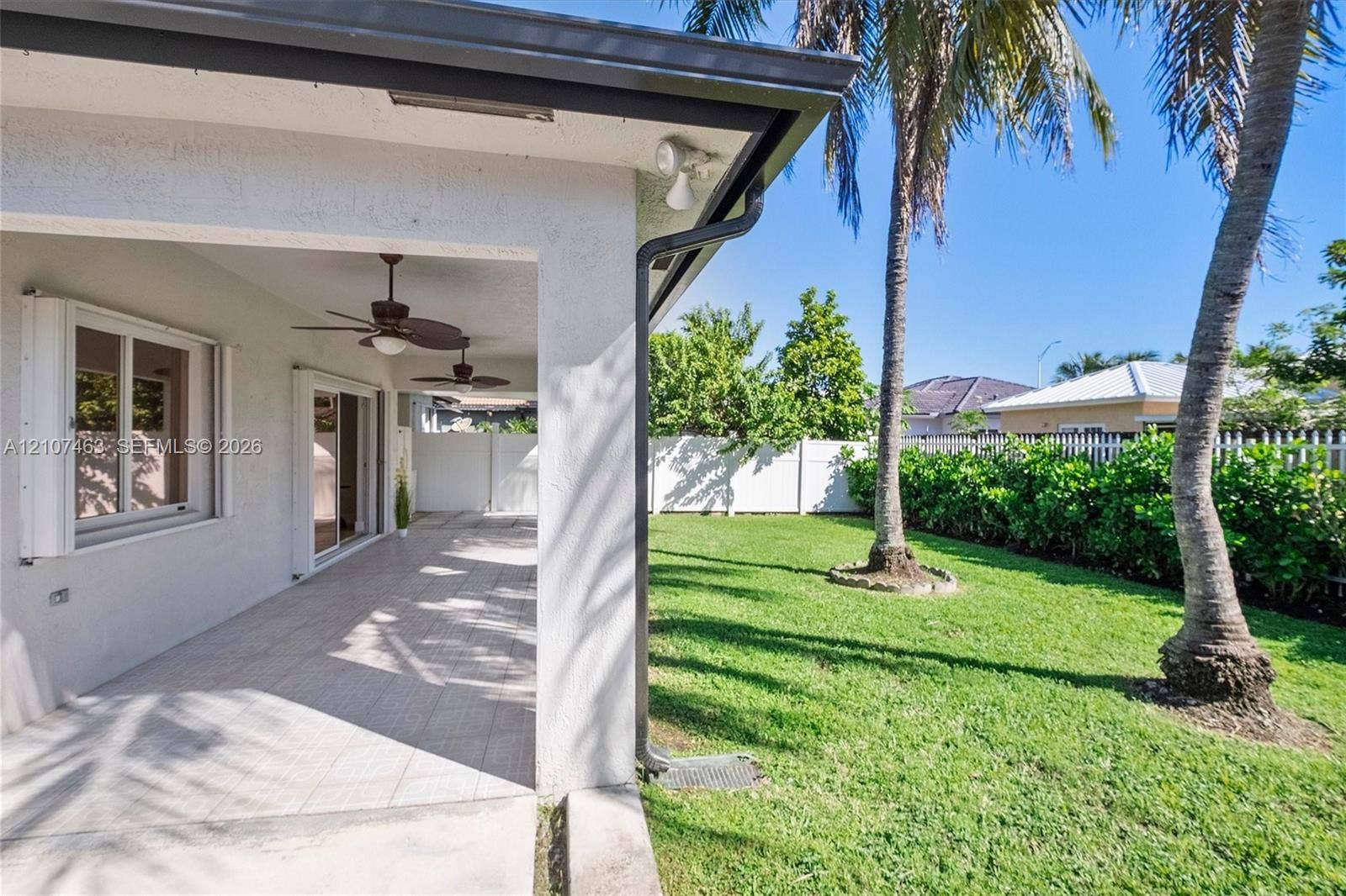 7071 W 6th Avenue, #N/A, Hialeah, Hialeah, FL 33014 Covered concrete patio featuring ceiling-mounted fans and sliding glass doors, overlooking a lawn enclosed by white vinyl fencing and palm trees