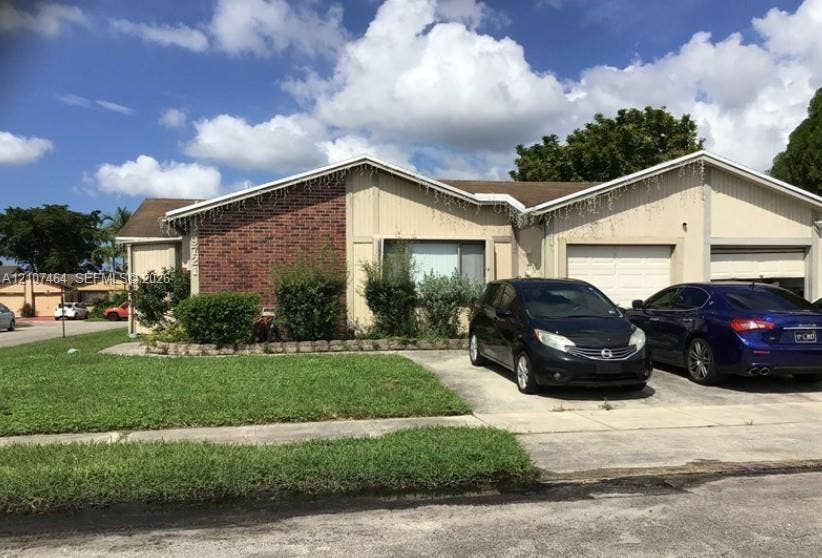 9721 W Daffodil Lane, Miramar, FL 33025 Single-story exterior featuring a brick facade, vertical wood-panel siding, dual-gabled roofline, two-car garage, and concrete driveway