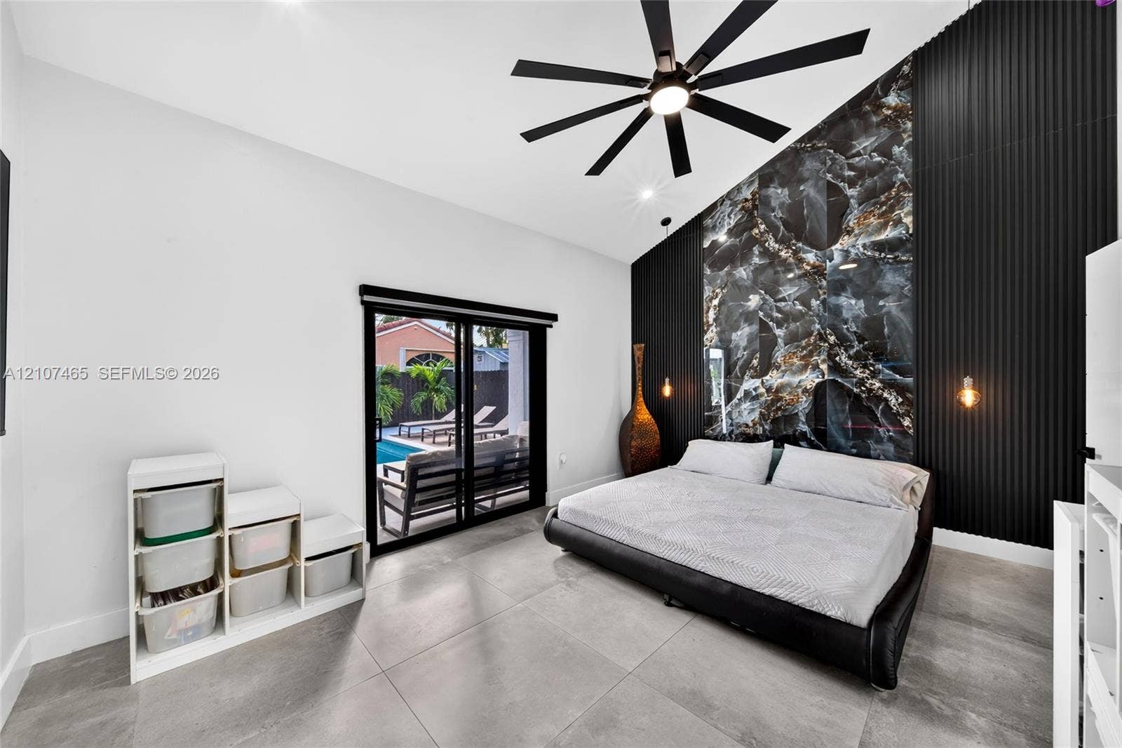 Vaulted ceiling room featuring large-format tile flooring, a statement accent wall with dark stone-finish cladding and vertical slats, a black multi-blade ceiling fan, and sliding glass doors