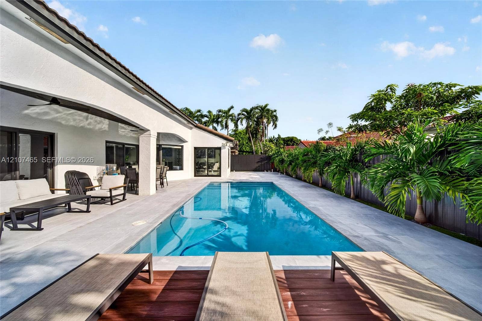 Rectangular in-ground pool featuring expansive tile decking, a covered patio area, and perimeter privacy fencing accented by mature tropical vegetation