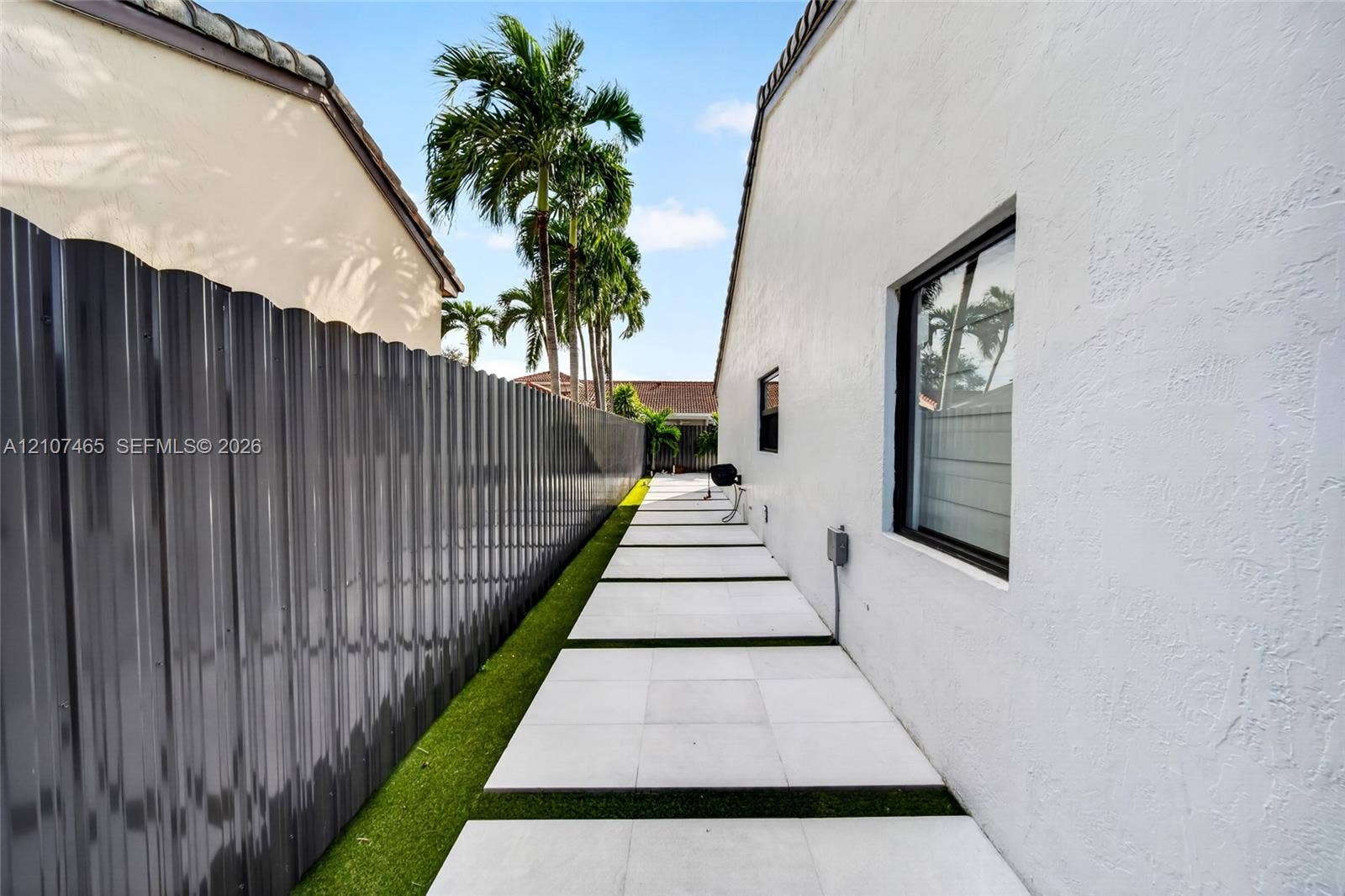 Side exterior featuring a white stucco facade, large-format tile pavers with turf inlay, and a corrugated metal privacy fence