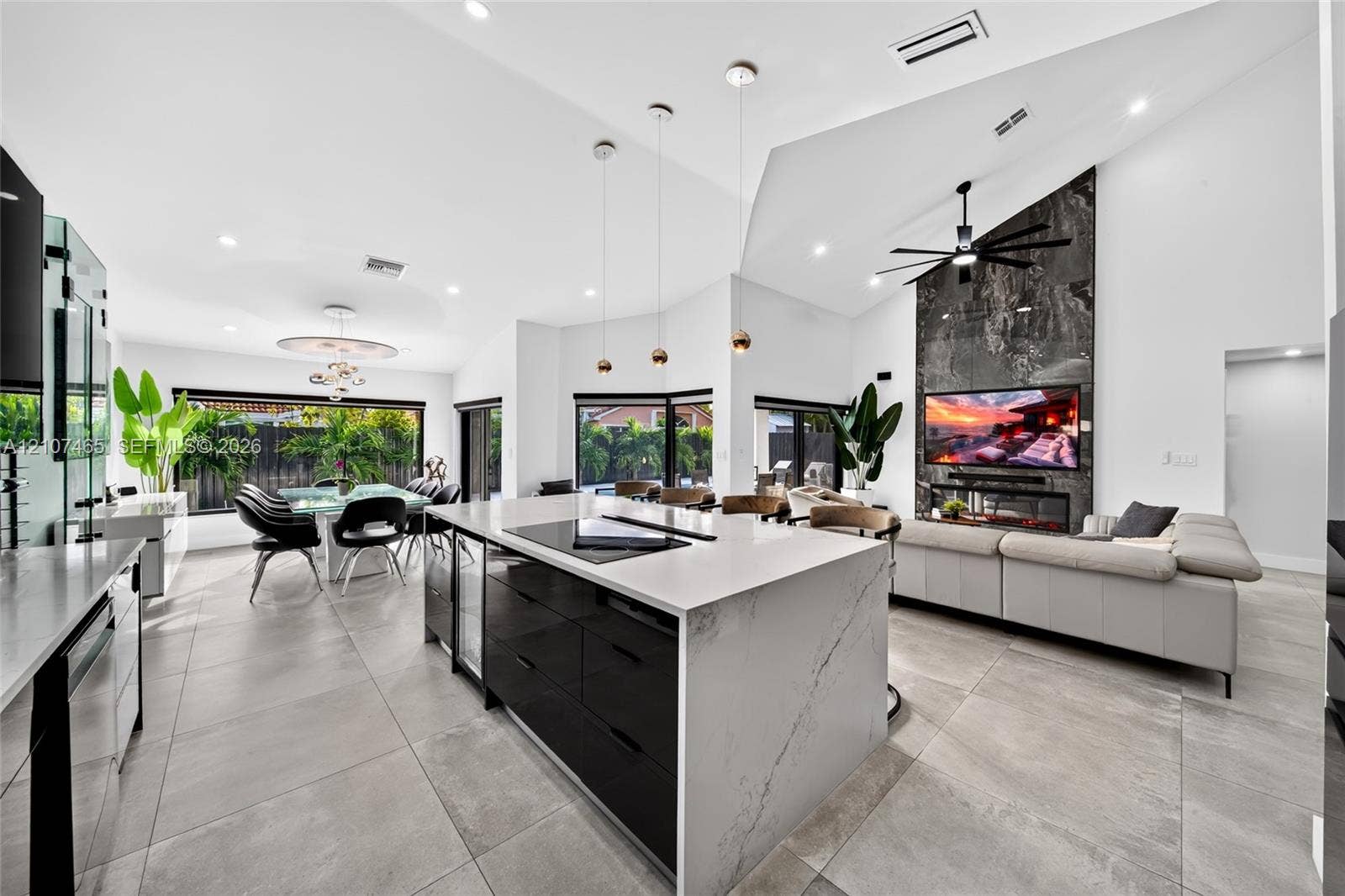 Open-concept floor plan featuring high vaulted ceilings, large-format tile flooring, a center island with stone-finish waterfall countertop, floor-to-ceiling stone-finish fireplace surround, and modern multi-light pendant fixtures