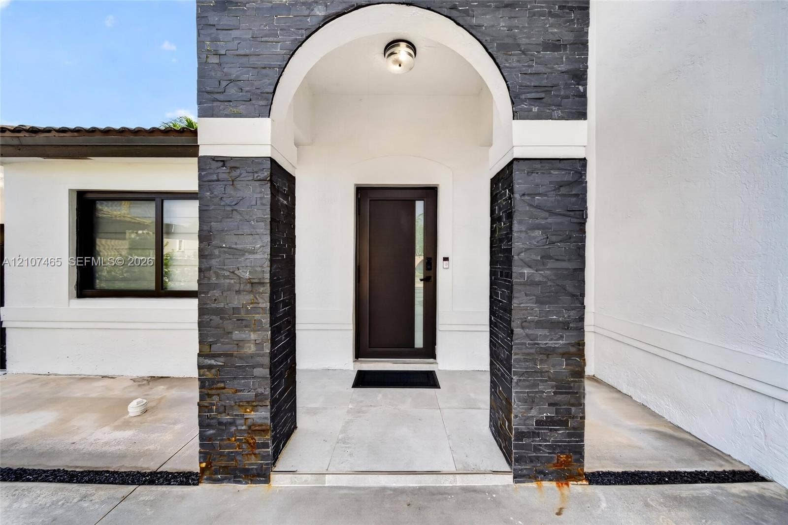 Covered entryway featuring a decorative arched portal, charcoal-toned stone veneer pillars, a dark-finished entry door with a vertical glass inset, and light-toned concrete surfacing