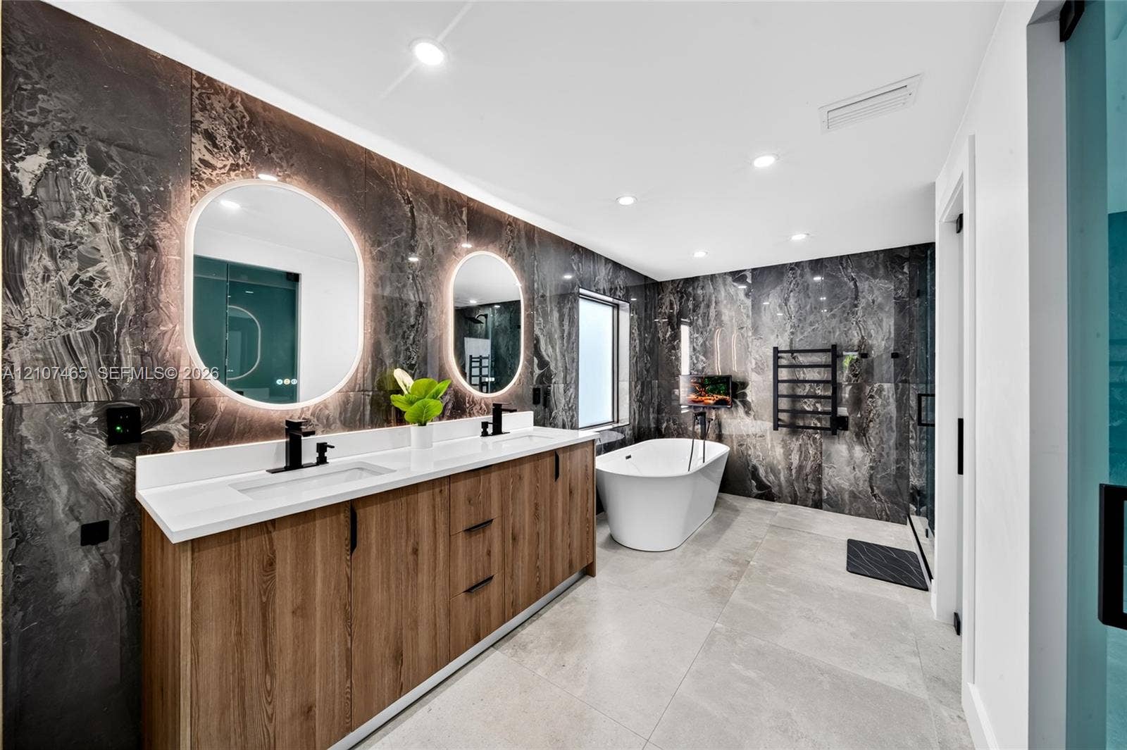 Dual-sink vanity with wood-finish cabinetry, stone-tiled accent walls, freestanding soaking tub, backlit oval mirrors, and matte black fixtures