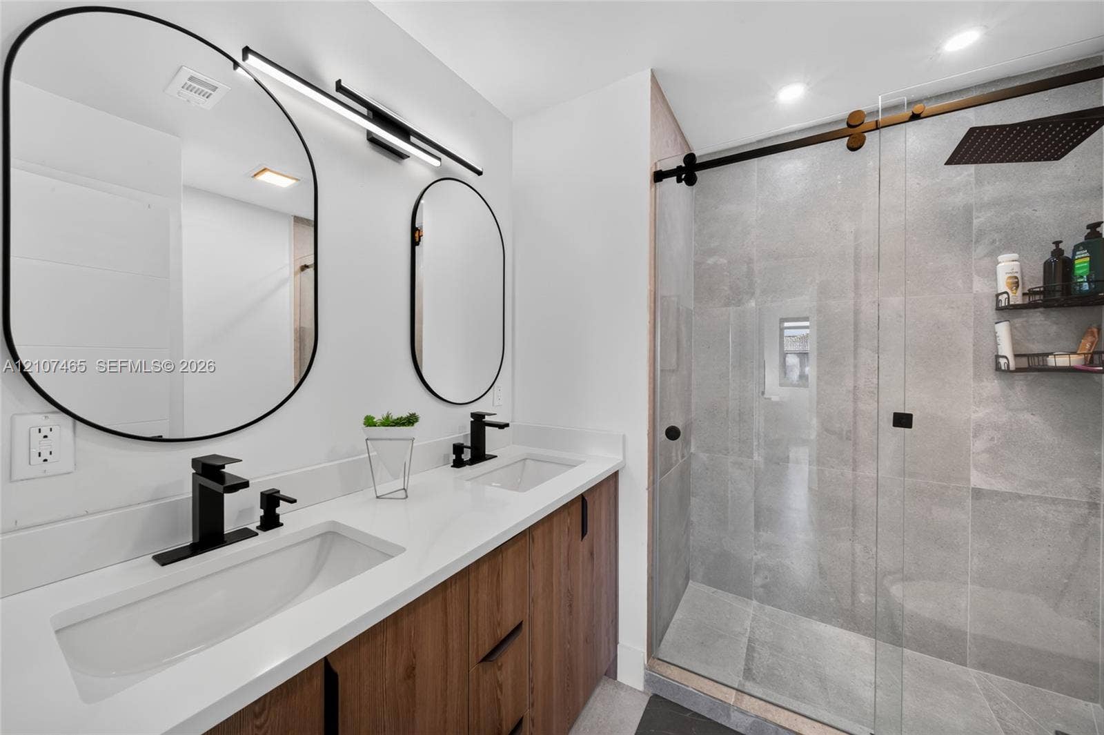 Dual-sink vanity with wood-finish cabinetry, quartz countertop, matte black fixtures, oval mirrors, and walk-in shower with floor-to-ceiling tile and glass sliding door
