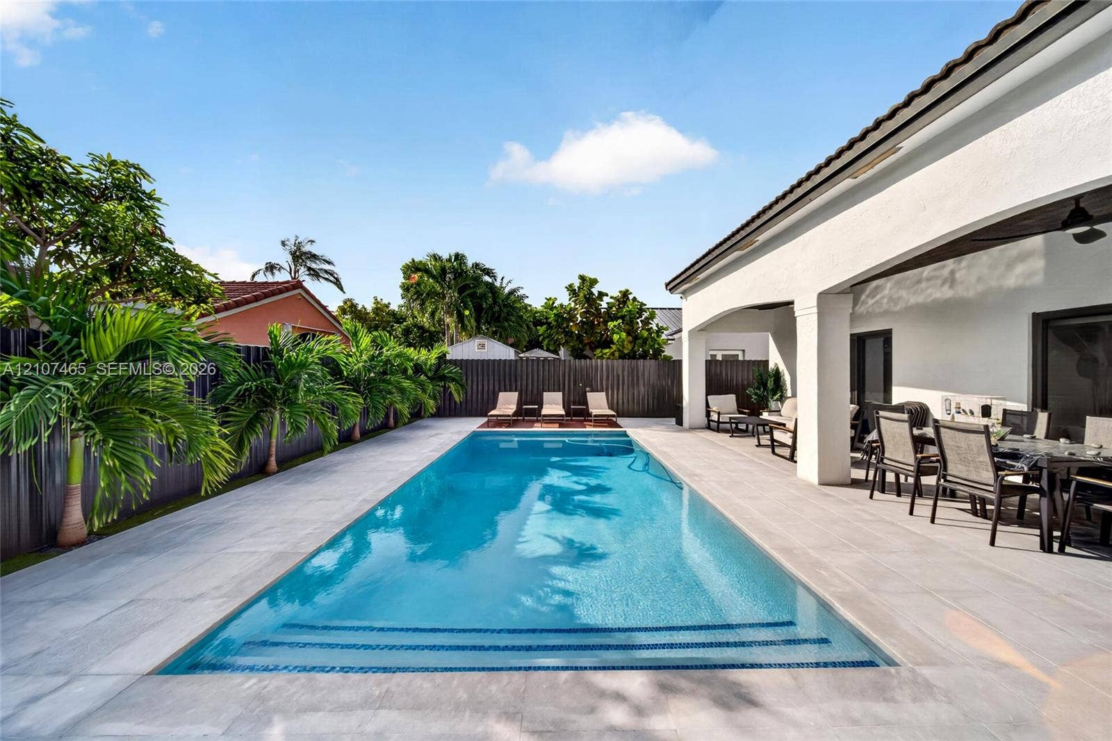 Rectangular in-ground swimming pool featuring a tiled waterline, integrated steps, and light-toned stone-finish paver patio surround, shaded by a covered veranda