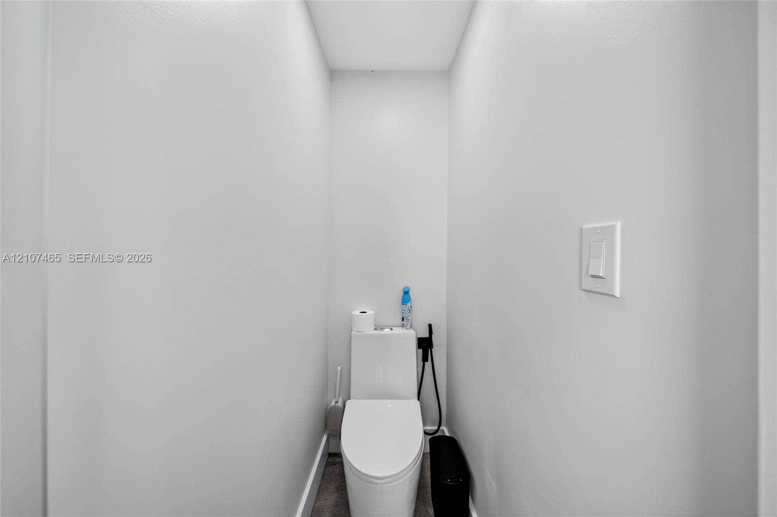 Compact restroom featuring a white dual-flush toilet, matte black bidet sprayer, and neutral-toned wall paint