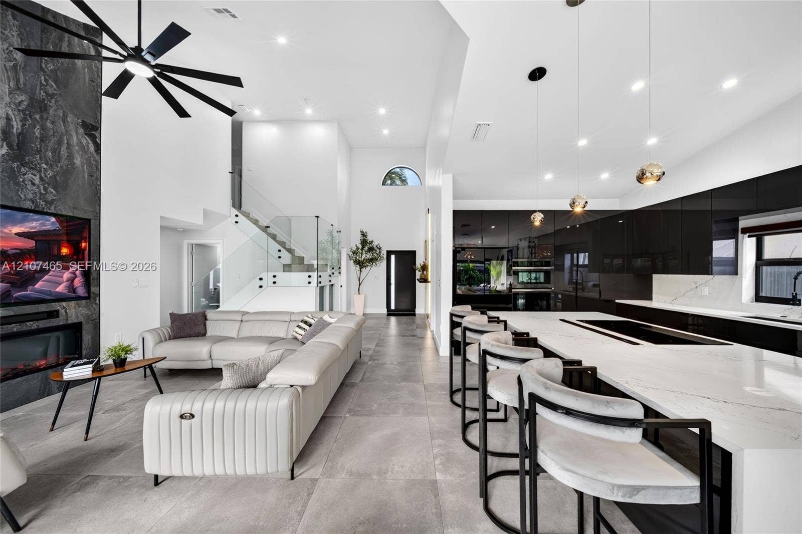 Open-concept living space featuring high ceilings, a full-height stone-clad fireplace, large-format tile flooring, a kitchen with high-gloss cabinetry, and a glass-paneled staircase