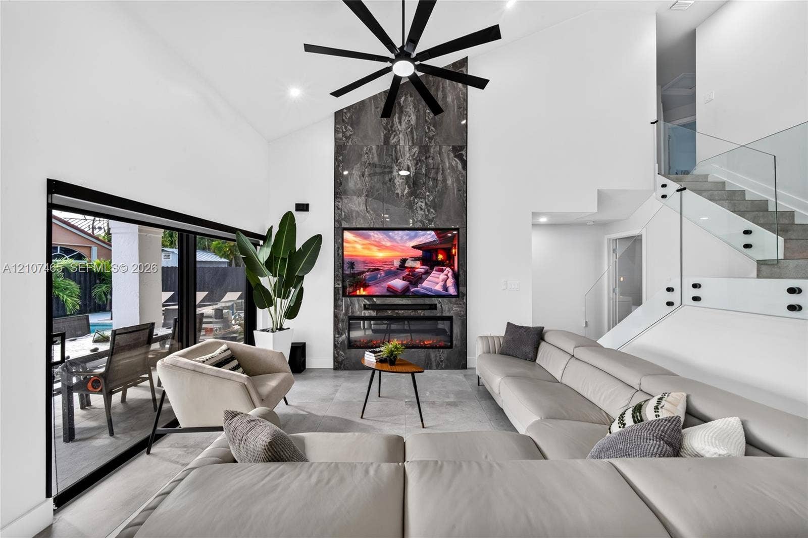 Vaulted ceiling with an oversized multi-blade ceiling fan, full-height stone-clad fireplace with integrated electric fire, large-format tile flooring, and a glass-paneled staircase railing