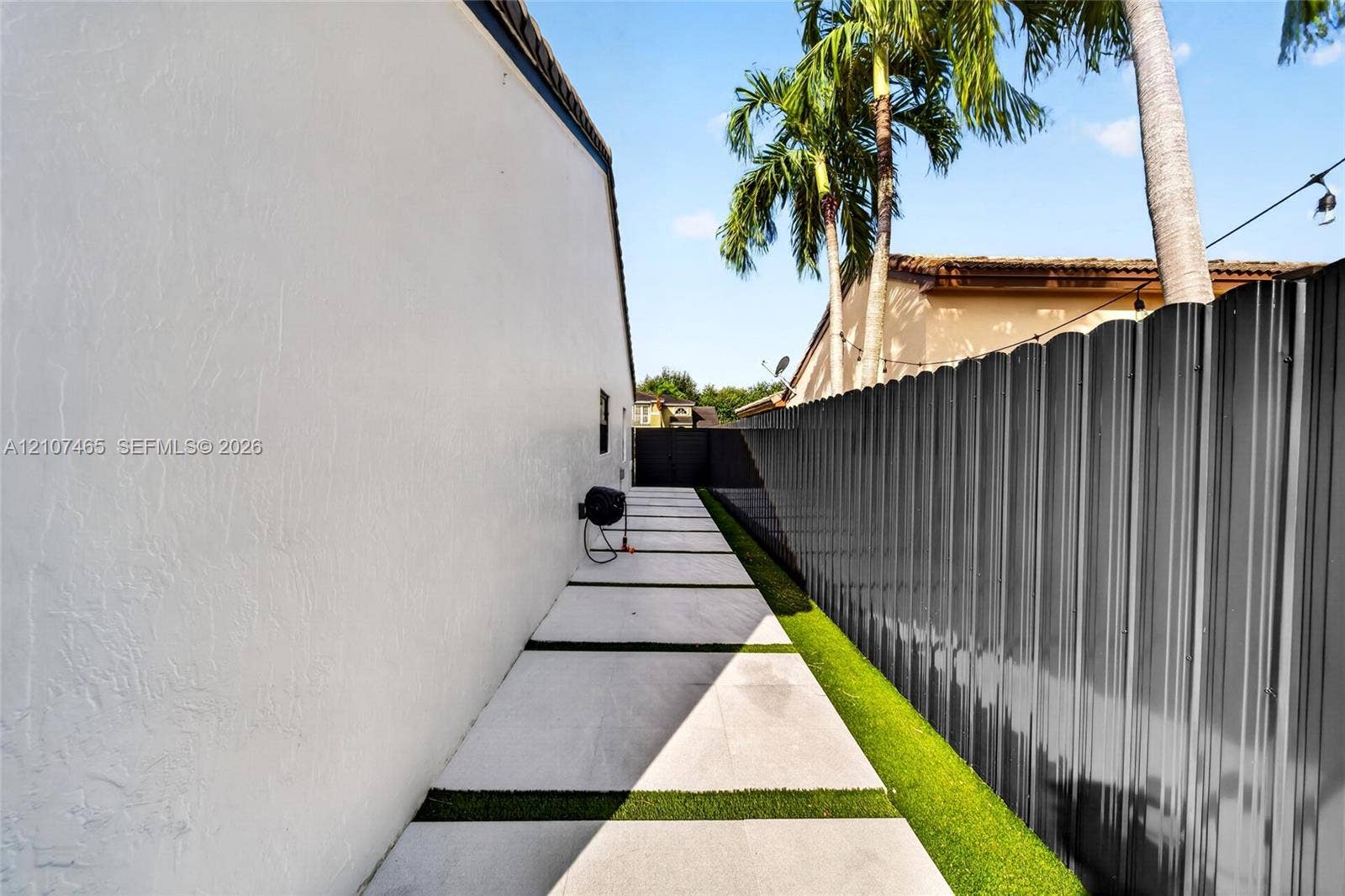 Side exterior walkway featuring oversized concrete pavers with turf inlays, white stucco exterior wall, and tall corrugated metal privacy fencing