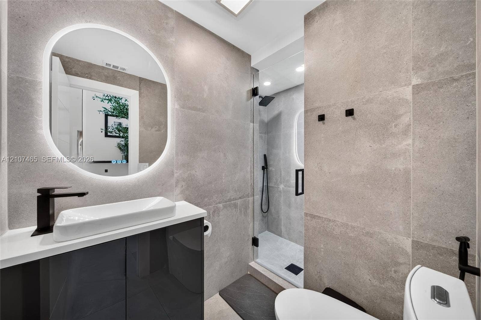 Modern bathroom featuring large-format grey tile wall cladding, a glossy charcoal vanity with a white vessel sink, a backlit oval mirror, and a walk-in shower with a glass enclosure and matte black fixtures