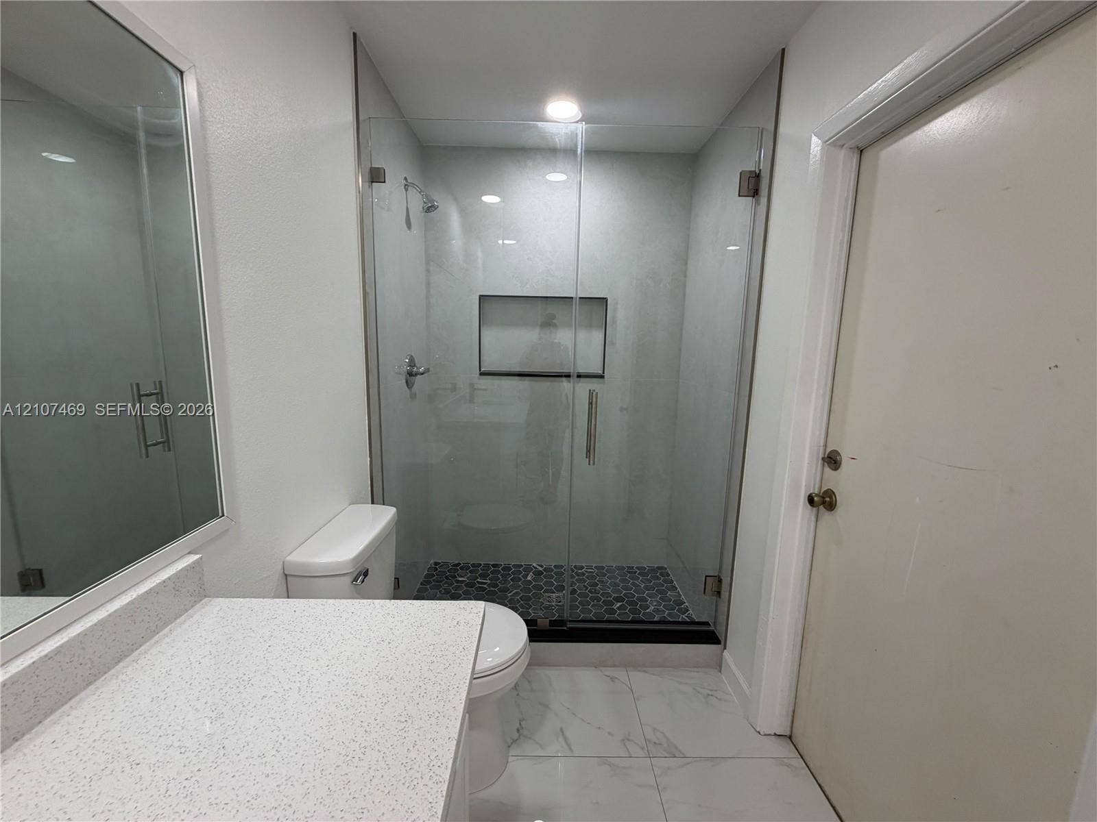 Bathroom featuring a glass-enclosed shower with a recessed wall niche, hexagonal tile shower flooring, large-format stone-patterned floor tile, and a vanity topped with a quartz countertop