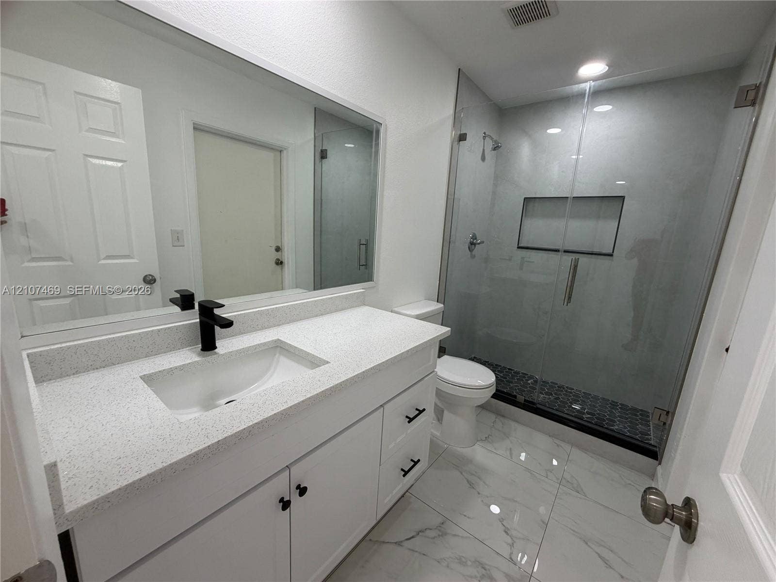 Bathroom featuring a white vanity with a light-toned solid surface countertop, matte black faucet, large wall-mounted mirror, glass-enclosed shower with a built-in recessed niche, and large-format tile flooring