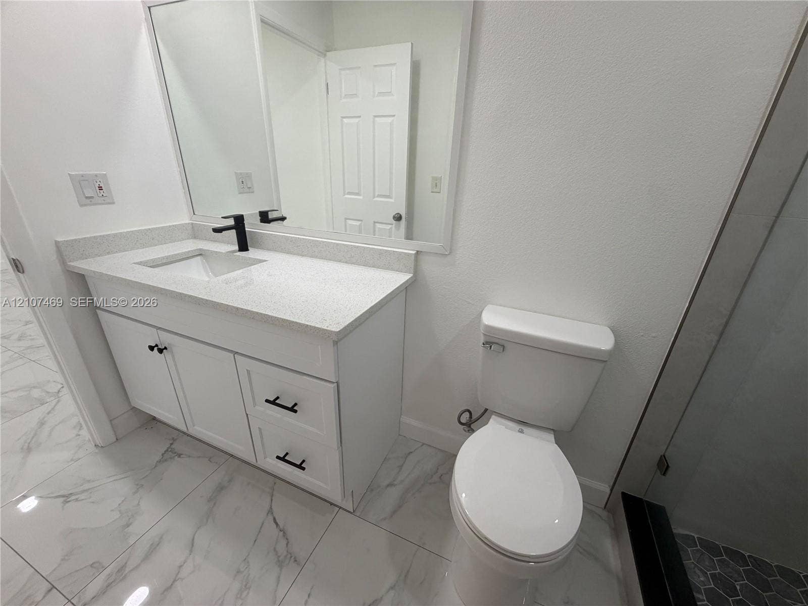 White vanity cabinet featuring a solid-surface countertop with an integrated basin, matte black hardware, a wall-to-wall mirror, and large-format porcelain tile flooring