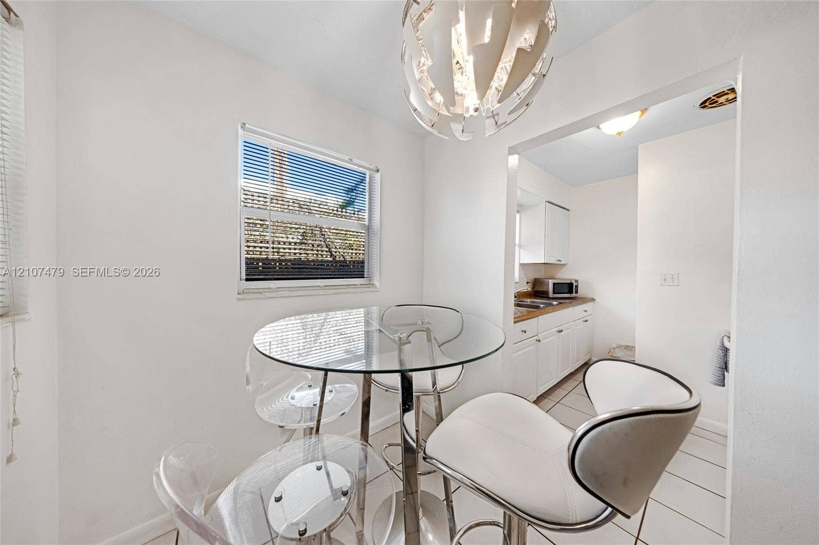 Dining area featuring a modern sculptural pendant light fixture, ceramic tile flooring, and a window with blinds, opening into a kitchen equipped with white cabinetry and stone countertops