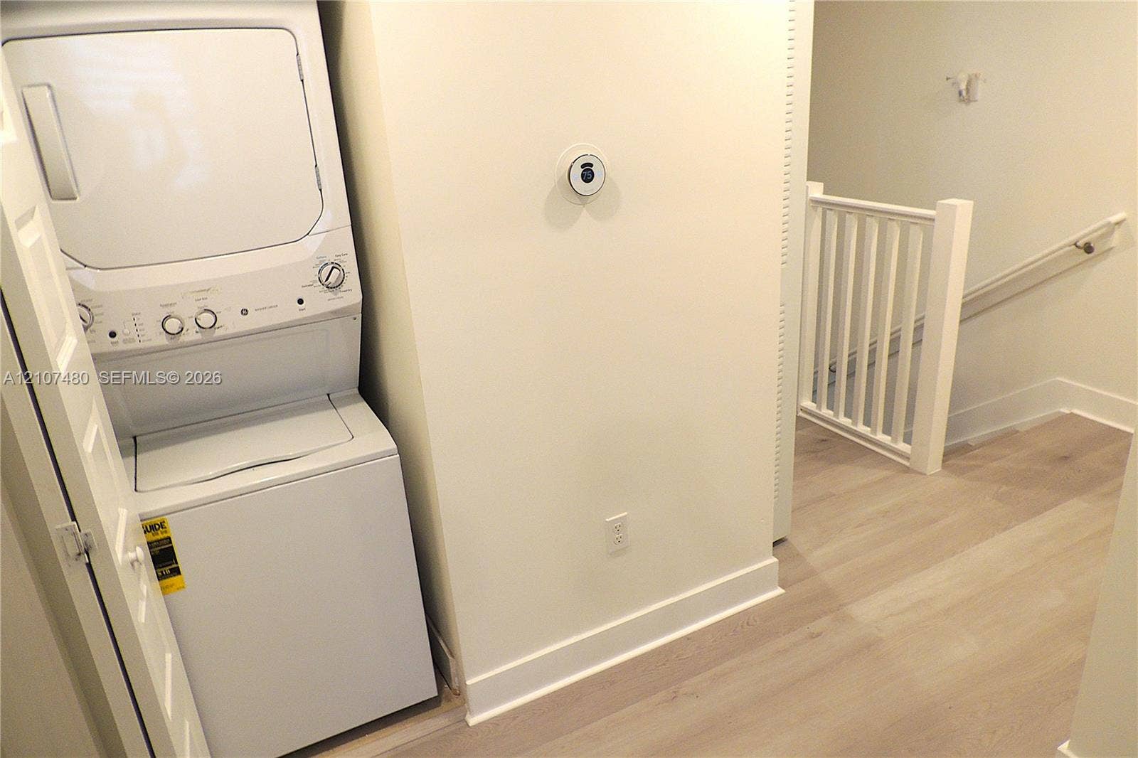 Laundry area featuring a stacked washer and dryer unit, wood-finish flooring, smart thermostat, and a white balustrade staircase