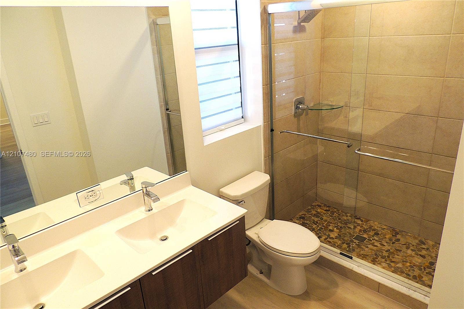 Double vanity with integrated sinks, quartz countertop, floating wood-finish cabinetry, walk-in shower with stone tile flooring and glass enclosure, and large wall-mounted mirror