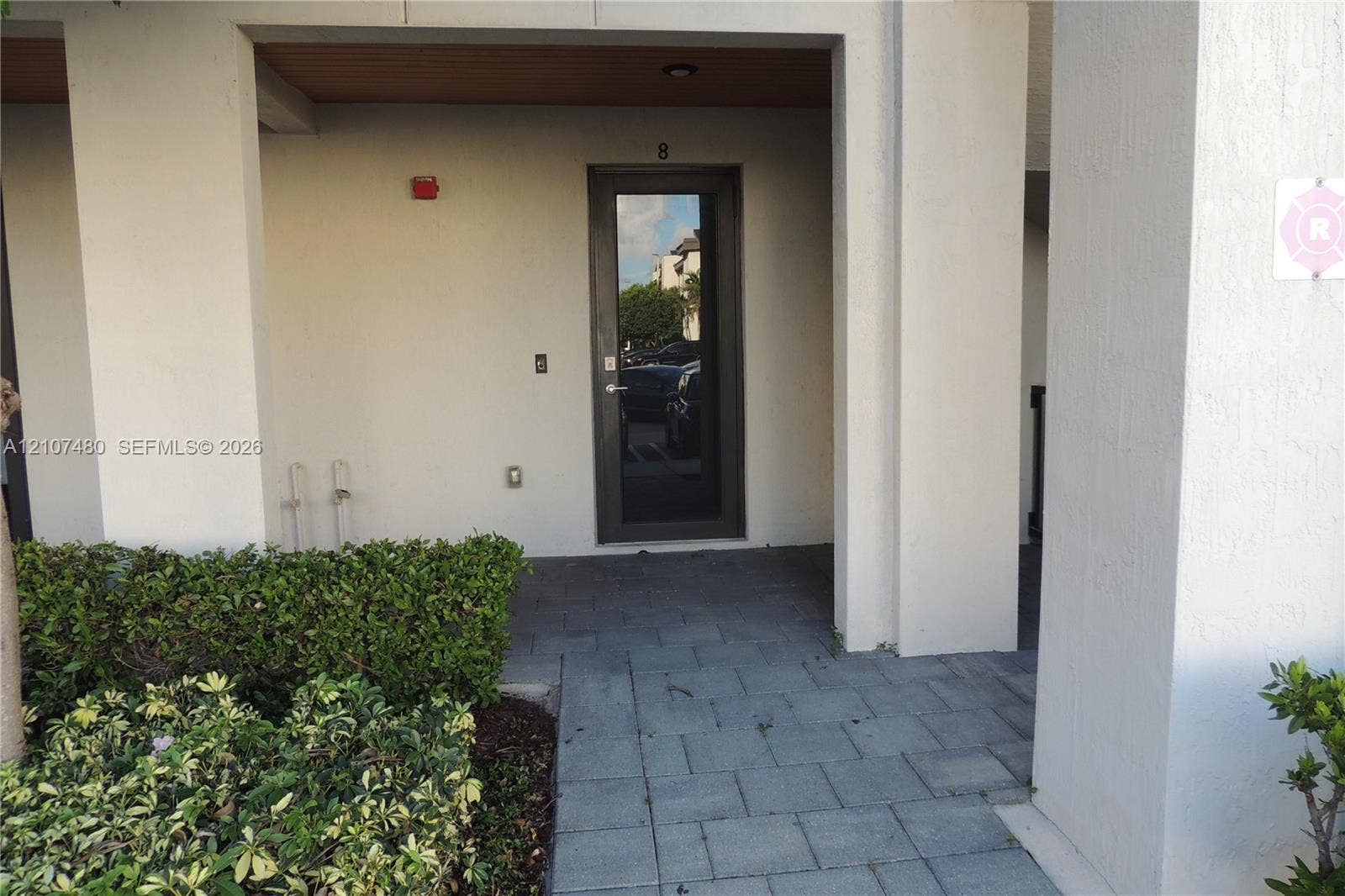 Covered entryway featuring a dark-framed glass door, exterior stucco finish, paver-stone walkway, and manicured shrubbery