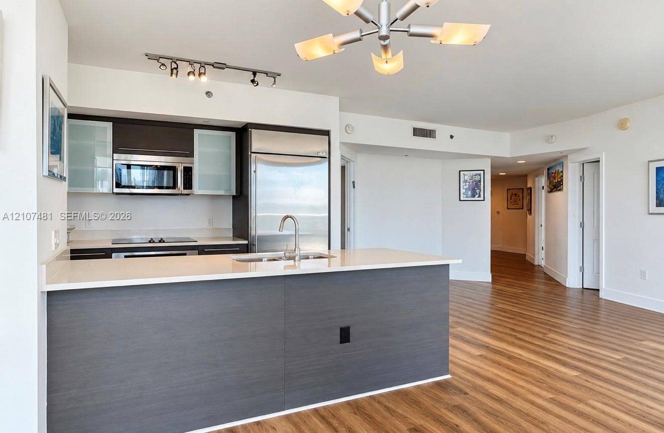 55 SE 6th Street, #2802, Miami, FL 33131 Kitchen featuring dark cabinetry with glass-panel accents, stone-surface countertops, stainless steel appliances, a central island with an integrated sink, and wood-finish flooring