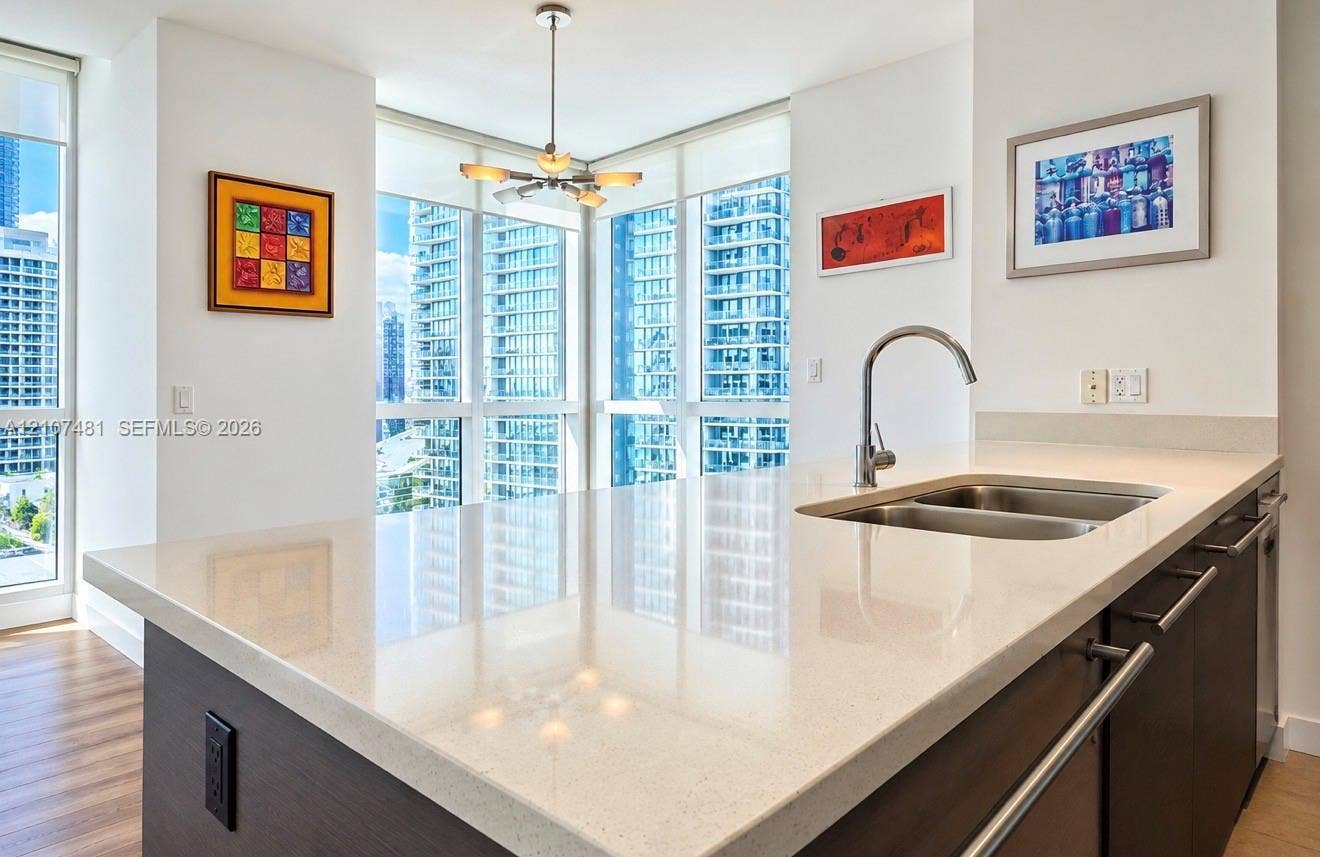 55 SE 6th Street, #2802, Miami, FL 33131 Kitchen island featuring a stone-surface countertop, double-basin stainless steel undermount sink, gooseneck faucet, dark-finish cabinetry, and floor-to-ceiling windows