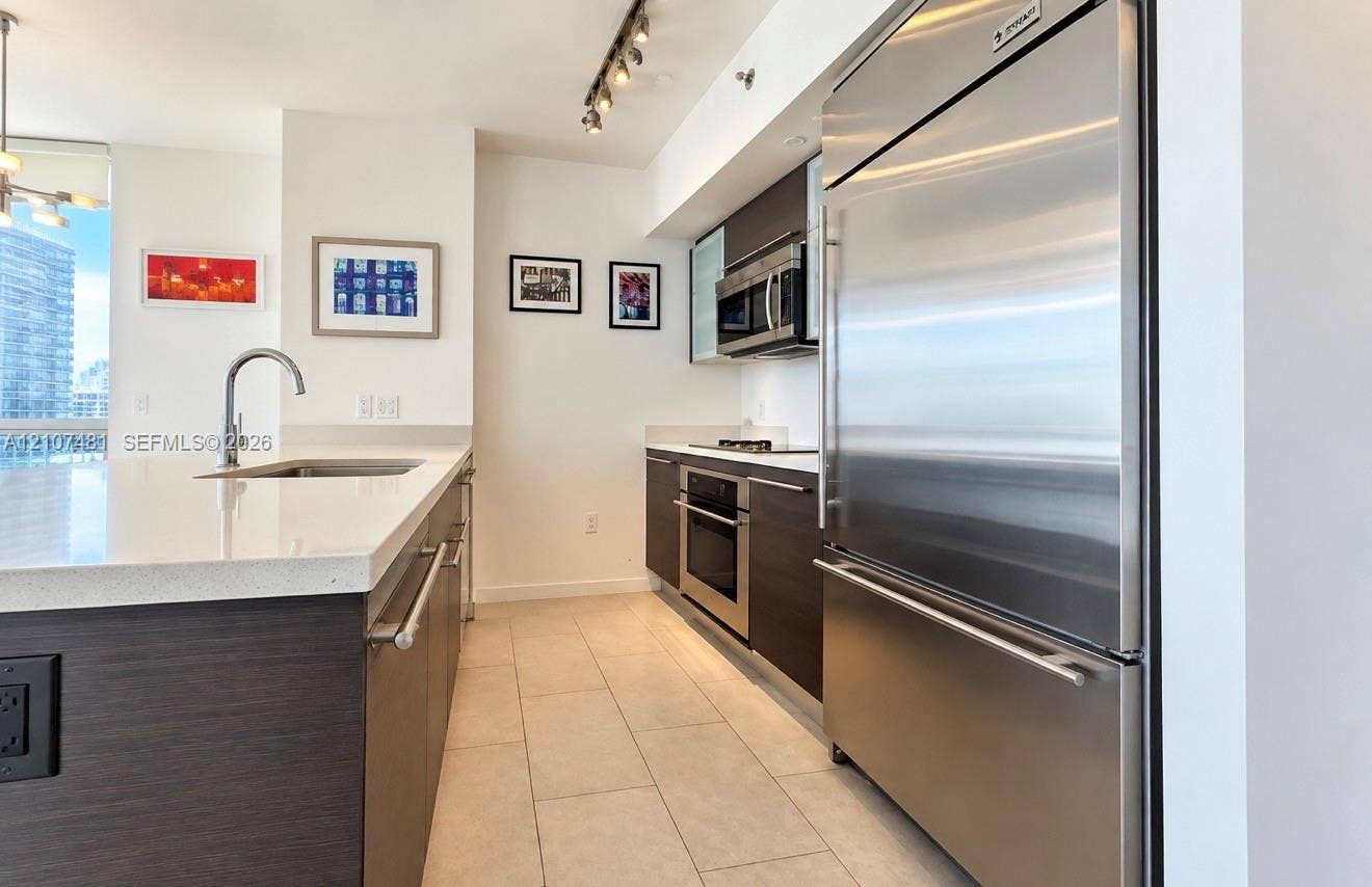 55 SE 6th Street, #2802, Miami, FL 33131 Kitchen featuring dark wood-finish cabinetry, stone-composite countertops, stainless steel appliances, a center island with an undermount sink, and tile flooring