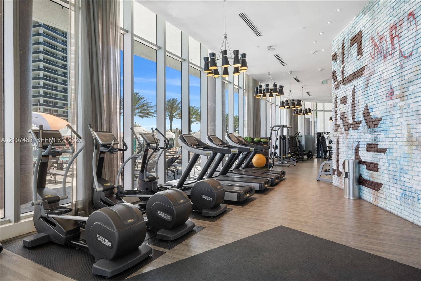 4111 S Ocean Drive, #2407, Hollywood, FL 33019 Fitness center featuring floor-to-ceiling windows, wood-finish flooring, decorative brick accent wall, and multi-tier modern chandelier lighting