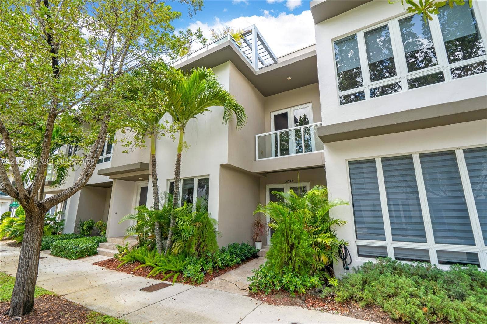 10545 NW 63rd Terrace, Doral, FL 33178 Modern multi-story exterior featuring a smooth stucco facade, expansive floor-to-ceiling windows, a private second-story balcony with glass railing, and a metal roof pergola