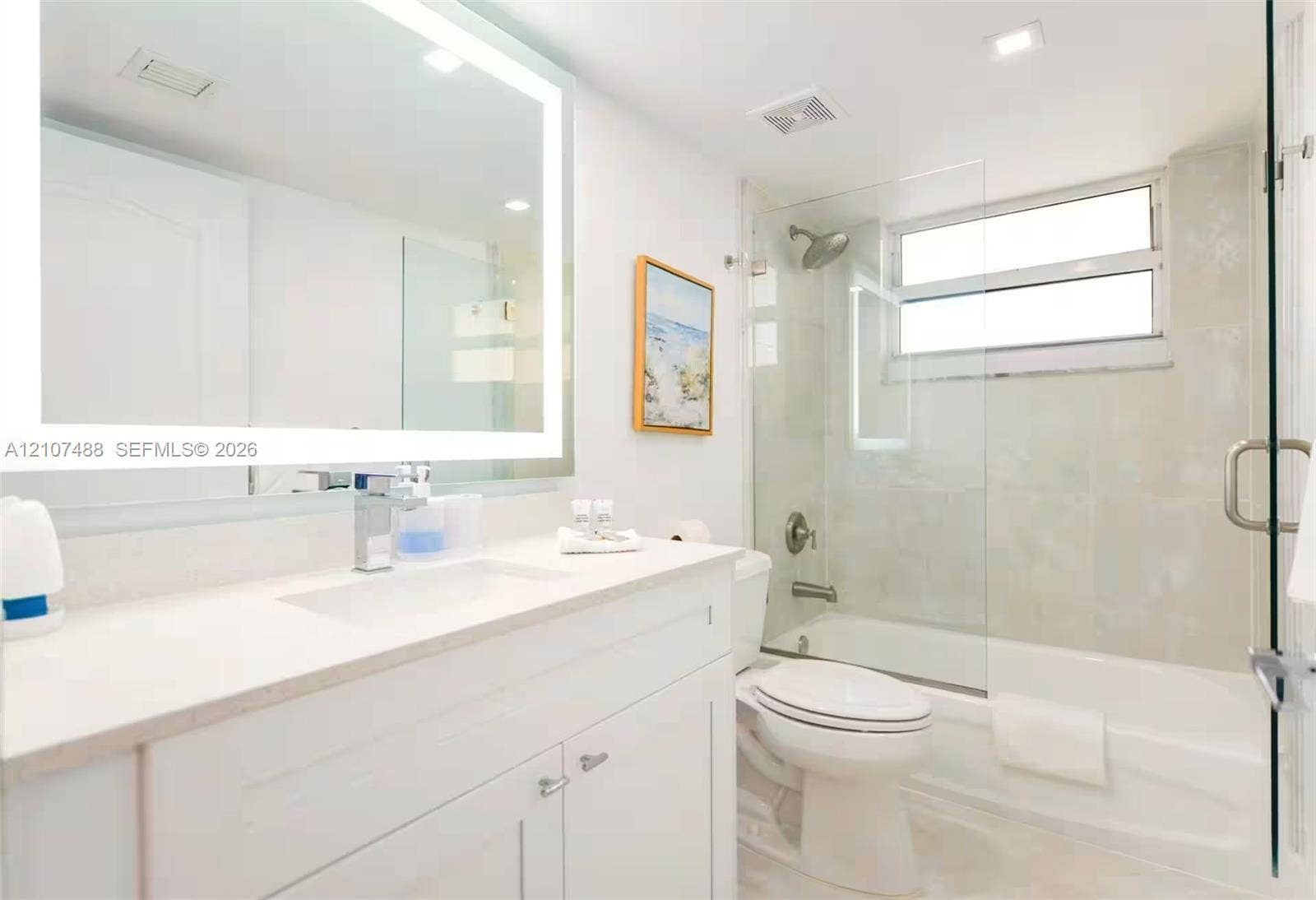 Bathroom featuring a white vanity cabinet with an integrated sink, backlit vanity mirror, glass-enclosed shower and tub combination with tile surround, and a horizontal window
