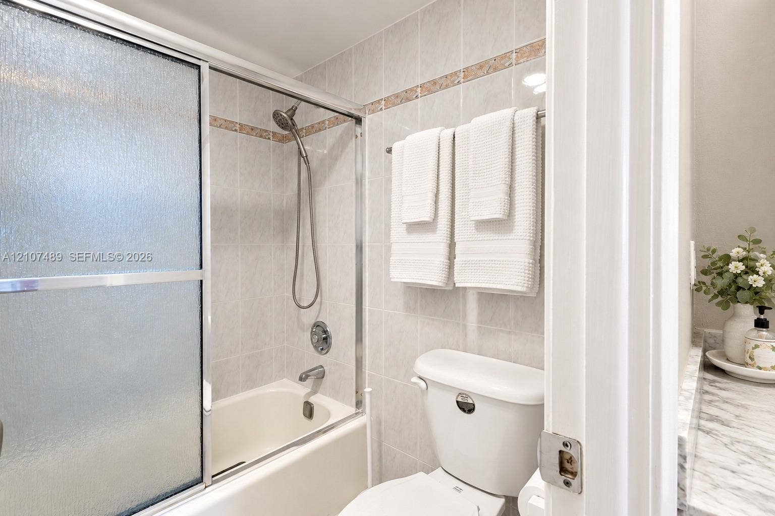 2000 S Ocean Boulevard, #9N, Lauderdale-By-The-Sea, Lauderdale By The Sea, FL 33062 Bathtub with glass sliding doors, integrated handheld shower fixture, light-toned wall tile with decorative accent border, and stone-finish vanity countertop