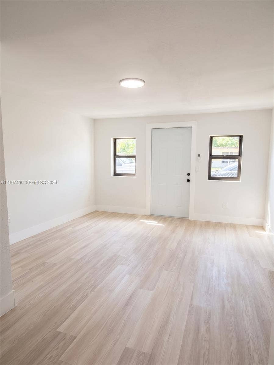 Spacious Interior space featuring light-toned wood-finish flooring, neutral wall paint, two windows with dark frames, and a solid-core entry door
