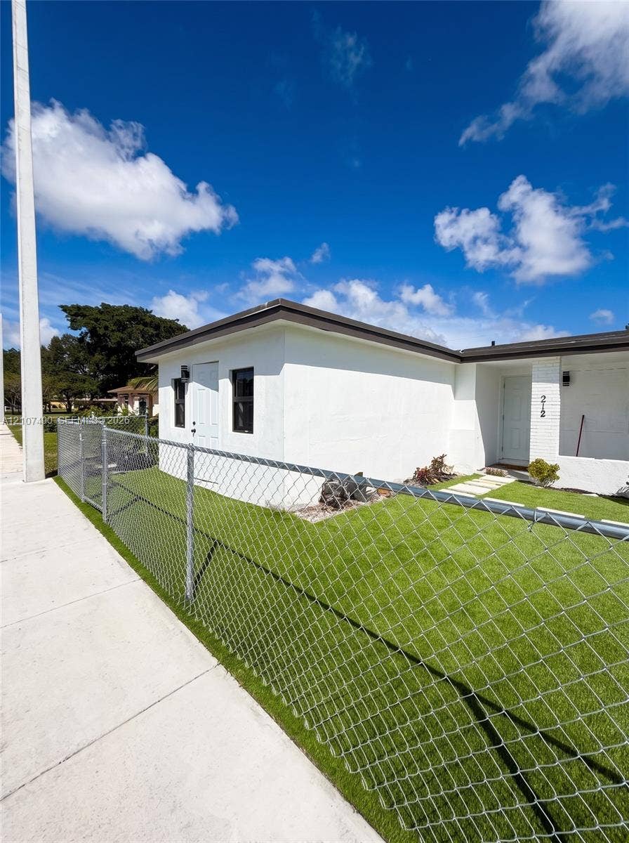 White stucco exterior featuring a flat roofline, chain-link perimeter fencing, and concrete walkway