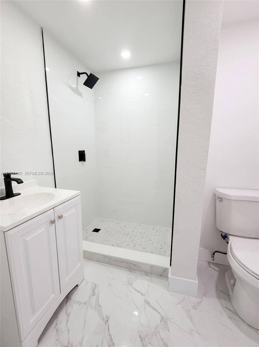 Walk-in shower with floor-to-ceiling white tile, glass enclosure, and matte black hardware, accompanied by a white vanity with integrated sink and stone-patterned flooring
