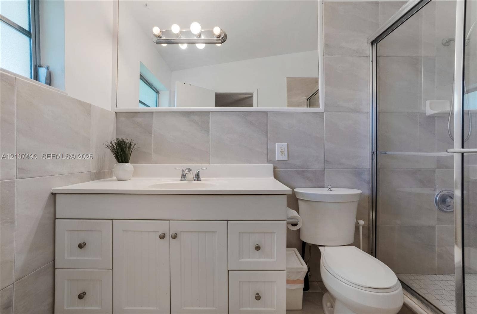 2645 NE 32nd Street, Fort Lauderdale, FL 33306 Bathroom featuring a white vanity cabinet, solid-surface countertop with integrated sink, large-format tile wall cladding, full-width mirror, and a glass-enclosed shower