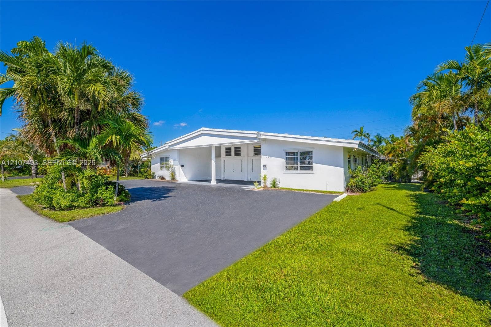 2645 NE 32nd Street, Fort Lauderdale, FL 33306 Single-story exterior featuring white stucco cladding, a paved driveway, a covered entrance, horizontal louvered windows, and mature palm trees