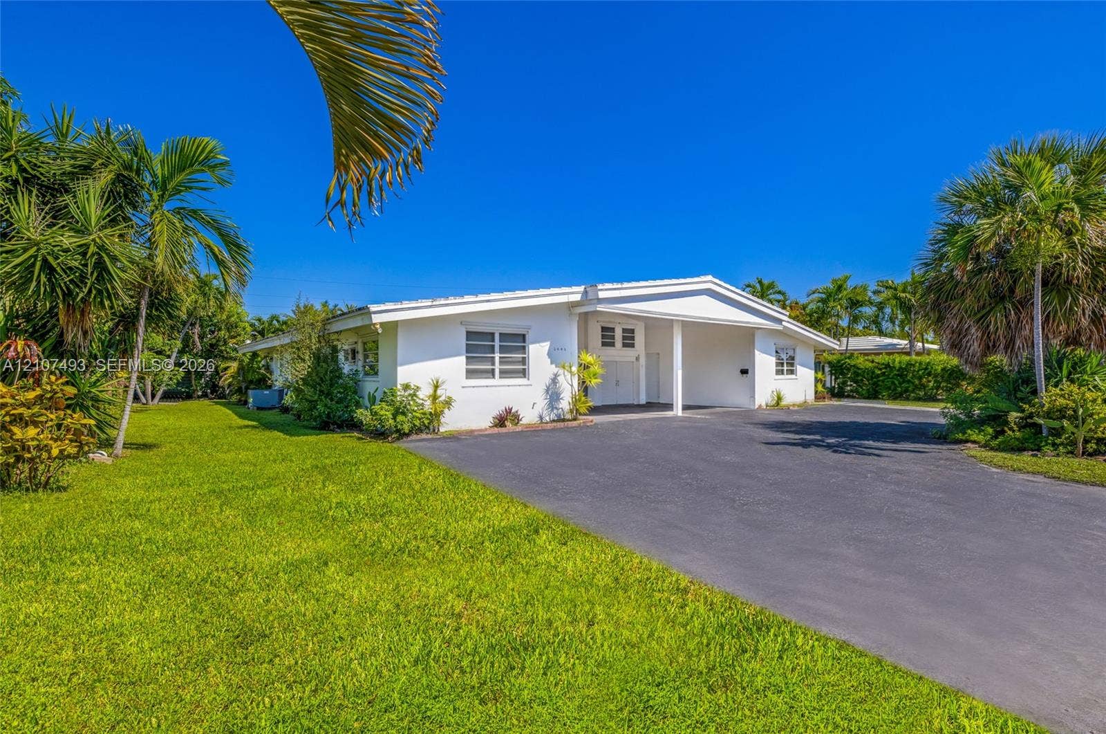 2645 NE 32nd Street, Fort Lauderdale, FL 33306 Single-story residence featuring white exterior cladding, a covered carport, and asphalt driveway, surrounded by mature palm trees and landscaped grounds