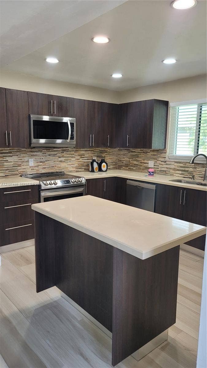 330 Racquet Club Road, #204, Weston, Weston, FL 33326 Kitchen featuring dark cabinetry, a central island with a solid-surface countertop, stainless steel appliances, a multi-tonal linear tile backsplash, and light-toned wood-finish flooring