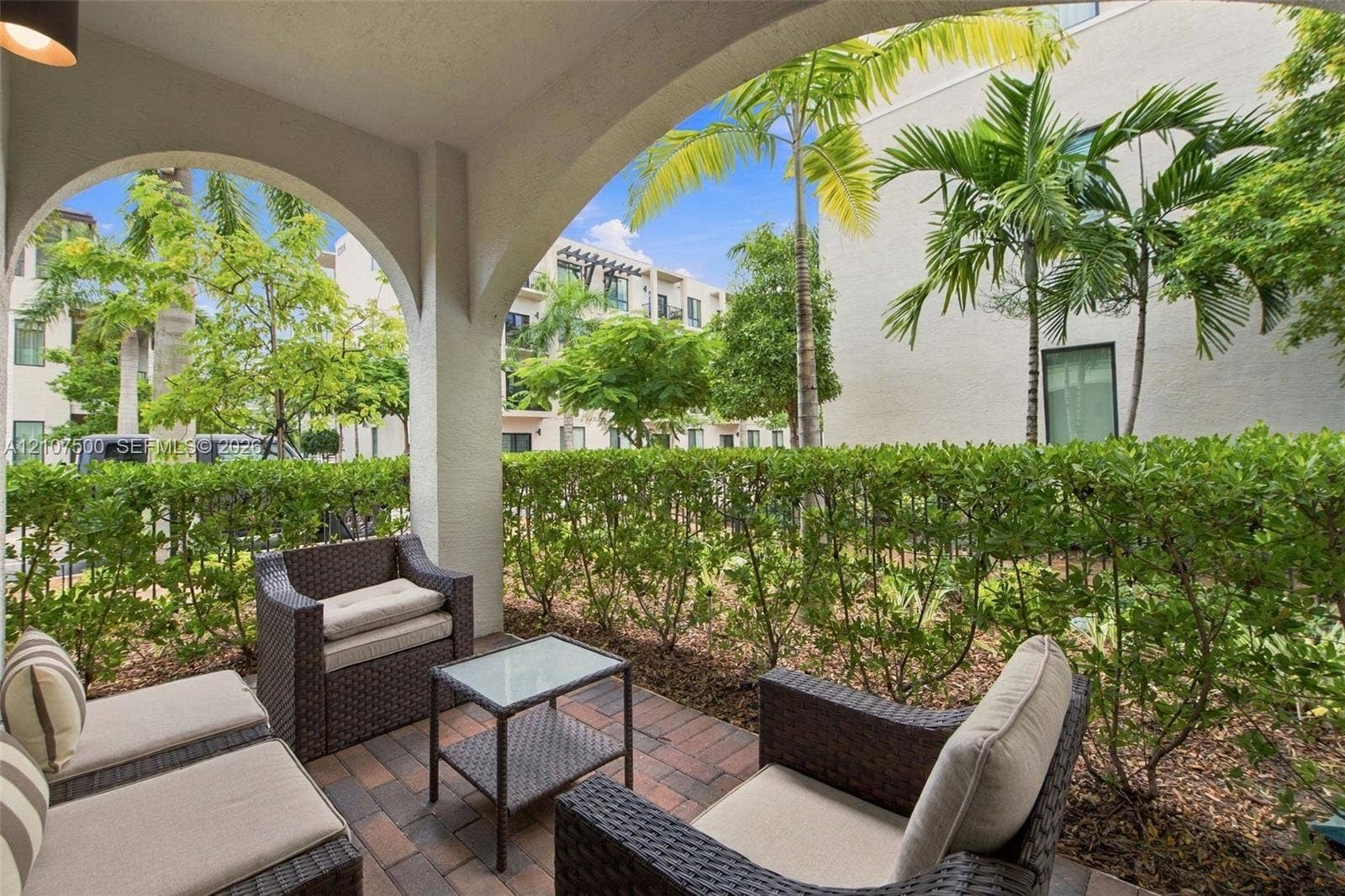 Covered patio featuring brick-paver flooring and architectural arched support columns, framed by mature hedges and palm trees