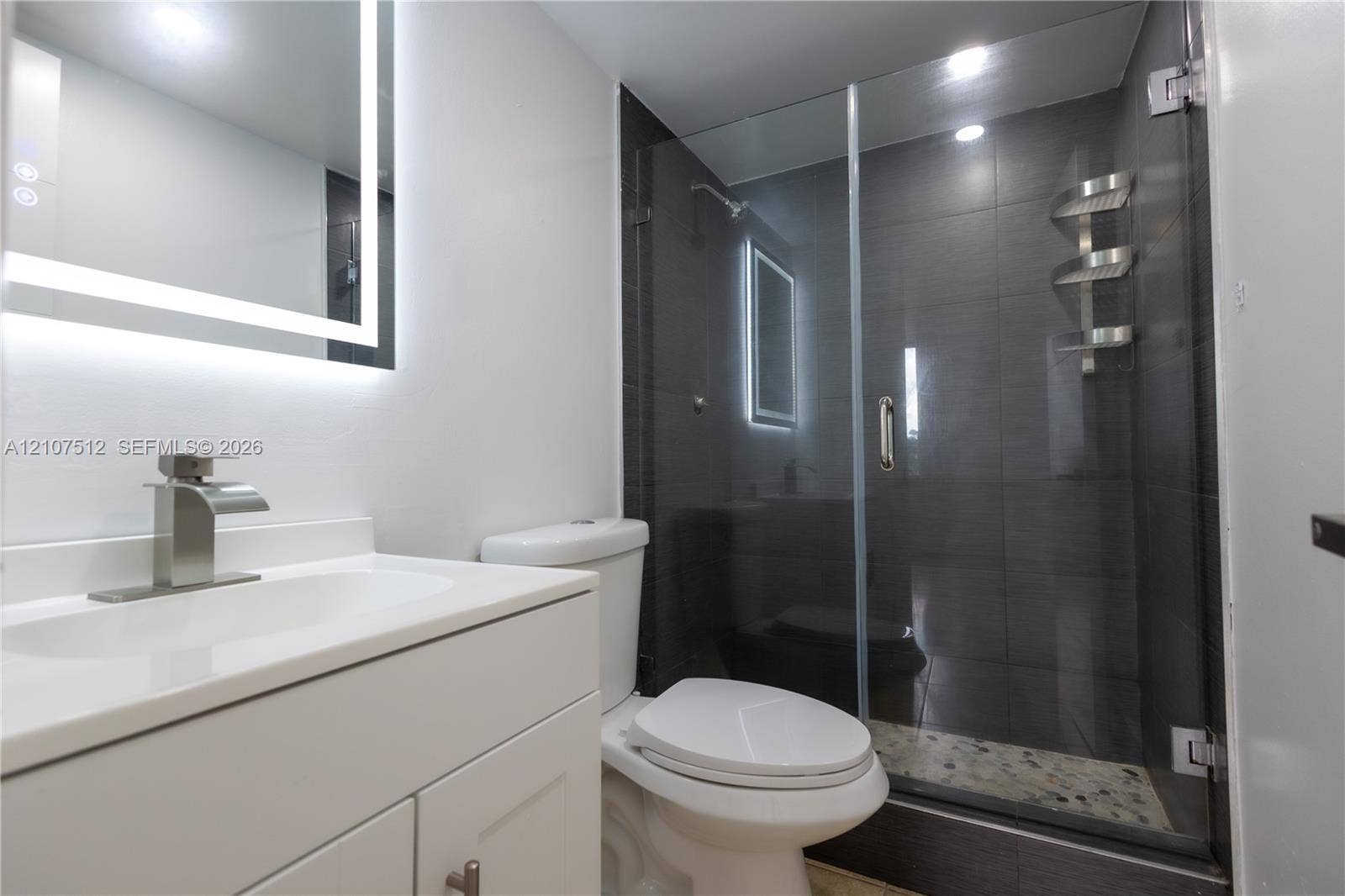1720 N River Drive, #302, Miami, FL 33125 Bathroom featuring a white vanity cabinet with an integrated countertop, a backlit LED vanity mirror, and a walk-in shower with dark tile surround, a glass door, and a pebble-stone shower floor